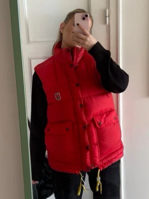 Fjällräven dunväst  - Snygg röd dunväst från Fjällräven med hög krage, tryckknappar och två stora fickor framtill. Klassisk logga på bröstet och justerbar dragsko nedtill. Perfekt för lager-på-lager och riktigt varm under kyliga dagar. Size m modell 167 lång ish 