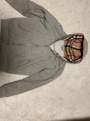 Grå zip hoodie från Burberry - Snygg grå zip hoodie från Burberry med den klassiska rutiga huvan i beige, svart, rött och vitt. Hoodien har dragkedja framtill, två fickor och ribbade muddar. Perfekt för dig som vill ha en stilren och exklusiv look.