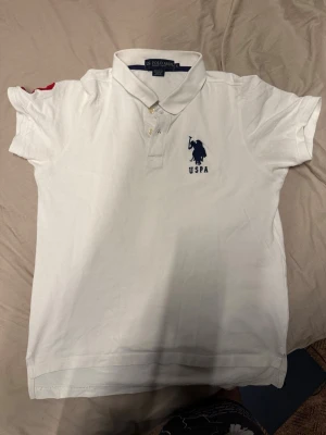 Vit pikétröja från U.S. Polo Assn - Klassisk vit pikétröja från U.S. Polo Assn med blå broderad logga och text på bröstet. Tröjan har krage, knappar framtill och korta ärmar. Perfekt till en avslappnad men ändå stilren look.