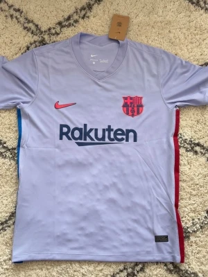 Barcelona bortatröja Nike ljuslila - Säljer en ljuslila FC Barcelona bortatröja från Nike med Rakuten- och UNICEF-logga. Tröjan har klubbmärke och Nike-logga i rött, samt blå och röda detaljer på sidorna. Tillverkad i lätt och ventilerande material, perfekt för fotboll eller träning.