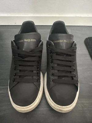 Svarta sneakers från Alexander McQueen - Stilrena svarta sneakers från Alexander McQueen med klassisk chunky sula och guldfärgad logga på plösen. Skorna har svarta skosnören och en vit, mönstrad yttersula med märkets logotyp undertill. Perfekta för dig som gillar exklusiv streetstyle.
