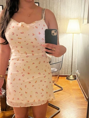 Vit blommig kortklänning med smala band - Söt vit kortklänning med små blommor i rosa och gult. Klänningen har smala axelband och en lätt figurnära passform. Perfekt för varma dagar och har en feminin, somrig look. Materialet känns lätt och luftigt.