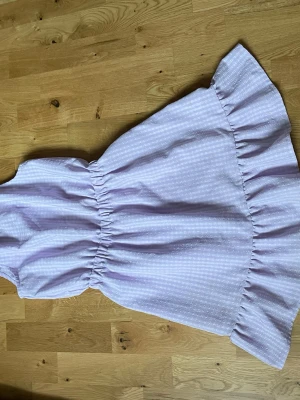 Ljuslila kortklänning med volang - Supersöt ljuslila kortklänning med diskret rutmönster och bred volangkant nedtill. Klänningen har resår i midjan för en snygg passform och är perfekt för varma dagar. Tillverkad i mjuk bomull som känns skön mot huden.