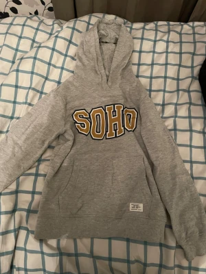 Grå hoodie med SOHO-tryck från 157 - Snygg grå hoodie från 157 med stort SOHO-tryck i gult och svart på bröstet. Klassisk känguruficka framtill och ribbade muddar vid ärmar och nederkant. Tillverkad i mjukt bomullsmaterial och har en skön huva.