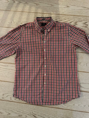 Rutig skjorta i rött och blått - Klassisk långärmad skjorta med rött, blått och vitt rutmönster. Skjortan har knappar hela vägen fram och en krage med knappdetaljer. Perfekt för dig som gillar en tidlös och avslappnad stil.