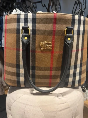 Burberry rutig handväska med läderdetaljer - Klassisk handväska från Burberry i beige, svart, rött och vitt rutmönster. Väskan har svarta läderhandtag, guldfärgade metalldetaljer och Burberrys logotyp framtill. Invändigt finns ett foder med Burberry-tryck och en innerficka med dragkedja.