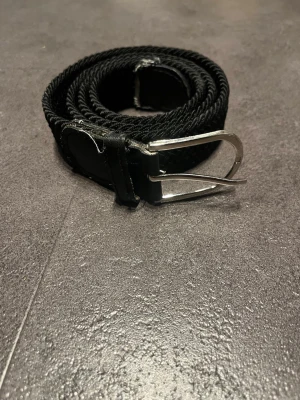 Svart flätat bälte med silverfärgat spänne - Snyggt svart flätat bälte med ett stilrent silverfärgat metallspänne. Bältet har detaljer i skinn vid spännet och ändarna, vilket ger en klassisk look. Perfekt för att lyfta vilken outfit som helst.