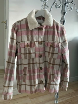 Rutig cardiganjacka med teddykrage - Snygg cardiganjacka från NLY trend i rutigt mönster med rosa, beige och bruna toner. Jackan har två bröstfickor med knappar, sidofickor och en mysig teddykrage. Perfekt för lager på lager och trendig look.