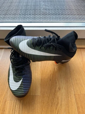 Nike Mercurial fotbollsskor svart/vit - Säljer ett par Nike Mercurial fotbollsskor med svart och vit färg, stora vita swoosh-loggor och tunna gröna och blå linjer. Skorna har snörning, strumpliknande skaft och dobbar i metall och plast för bra grepp på planen. Perfekta för dig som vill ha stil och funktion på fotbollsplanen. Finns bara några i världen 