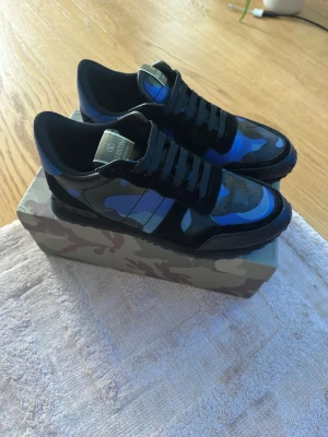 Valentino blå camo sneakers - Snygga sneakers från Valentino med blå och svart camo-mönster. Skorna har svarta detaljer, svarta skosnören och en klassisk låg siluett. Materialmix av textil och mocka ger en exklusiv känsla. Perfekta för dig som vill sticka ut med stil. OBS!!! Endast använda 1 gång 