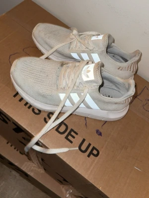 Beige Adidas sneakers med vita ränder - Säljer ett par snygga vita sneakers från Adidas med klassiska vita ränder på sidorna. Skorna har en stickad ovandel i textil och vita detaljer på plösen och sulan. Perfekta för dig som gillar bekväma sneakers med modern look. Storlek 40 använt några gånger finns lite smuts fläckar som går bort 