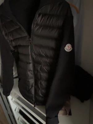 Svart cardigan från Moncler - Snygg svart pufferjacka från Moncler med quiltad front och stickade ärmar. Jackan har dragkedja framtill och Moncler-logga på ena ärmen. Perfekt för kalla dagar och ger en stilren look.