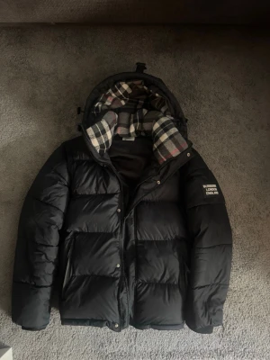 Svart pufferjacka från Burberry - Säljer en svart pufferjacka från Burberry med klassiskt rutigt foder i huvan. Jackan kan även göras till en riktig fet väst som är perfekt till våren som du ser på bilden, jackan är princip ny skick inga skavanker. Pris kan även diskuteras