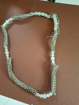 Halsband med vita pärlor och kedja - Unikt halsband med vita runda pärlor och grov silverfärgad kedja. Pärlorna är jämnt placerade längs halsbandet och ger en cool kontrast till den metalliska kedjan. Perfekt för dig som gillar att sticka ut med accessoarerna.