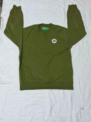 Olivgrön sweatshirt från Benetton - Olivgrön sweatshirt från United Colors of Benetton med rund halsringning och ribbade muddar. Tröjan har en liten patch med logga på bröstet och är tillverkad i mjuk bomull. Perfekt för en avslappnad och stilren look.