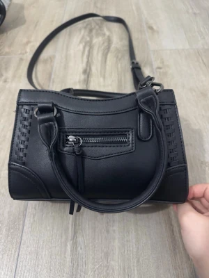 Svart handväska med axelrem - Snygg svart handväska i konstläder med både handtag och justerbar axelrem. Väskan har en liten dragkedjeficka framtill och dekorativa sömmar på sidorna. Perfekt storlek för det viktigaste och enkel att matcha till olika outfits.