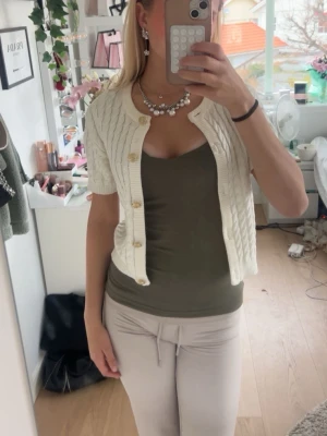 Kortärmad kabelstickad kofta, vit - Säljer en söt kortärmad kofta i kabelstickad modell med rund hals och stora dekorativa knappar. Koftan är vit och har en croppad passform som passar perfekt till linne eller topp under. Materialet känns mjukt och bekvämt, perfekt för lager på lager.