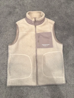 fleeceväst - Mysig beige fleeceväst från Jack & Jones Originals Studio med hög krage och dragkedja framtill. Västen har en kontrasterande bröstficka i ljusgrått med dragkedja och två stora fickor framtill. Perfekt för lager-på-lager och chill stil.