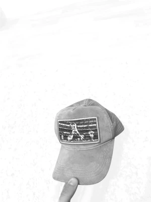 Grå meshkeps med boxningsmotiv - Säljer en grå keps med mesh på sidorna och bak, samt ett broderat motiv framtill som visar en boxningsscen. Kepsen har böjd skärm och justerbar rem bak. Perfekt för dig som gillar sportig och avslappnad stil.