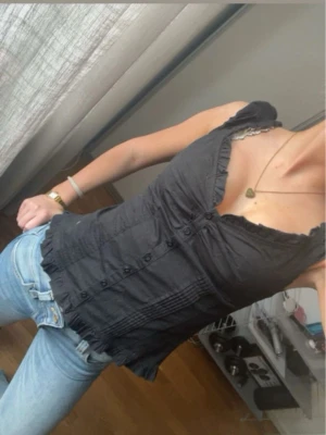 Svart blus med volang och knappar - Snygg svart blus med volangkant vid halsringning och nederkant. Modellen är figurnära med knappar framtill och fina sömdetaljer. Perfekt till jeans eller kjol för en trendig look.