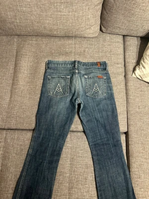 Blå bootcut jeans från 7 For All Mankind - Säljer ett par klassiska blå jeans från 7 For All Mankind med bootcut-modell. Jeansen har tydliga kontrastsömmar och ikonisk A-brodering på bakfickorna. Tillverkade i mjuk denim med snygg tvätt och normal midja. Perfekta för en avslappnad och trendig look.
