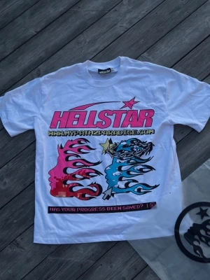 Vit Hellstar graphic t-shirt - Säljer en vit t-shirt från Hellstar med stort färgglatt tryck framtill. Motivet har rosa och blå flames, ansikten och texten 'HELLSTAR' i rosa samt coola detaljer. Klassisk passform och rund hals. Perfekt för dig som gillar streetwear och statement-plagg.