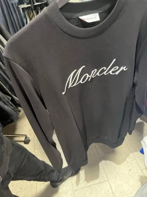 Svart Moncler sweatshirt med logga - Svart sweatshirt från Moncler med stor vit broderad Moncler-text på bröstet och klassisk rund hals. På ärmen sitter ett tygmärke med Monclers logga. Tröjan har långa ärmar och ribbade muddar. Perfekt för en clean och stilren look. Äkta prislapp kommer med för mer bilder skriv privat 