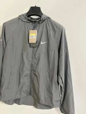 Grå vindjacka Nike  - Snygg grå vindjacka från Nike med dragkedja framtill och justerbar huva. Jackan har två sidofickor och en vit Nike-logga på bröstet. Tillverkad i lätt och tunt material som passar perfekt för blåsiga dagar.