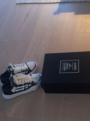 AMIRI x YASUTO sneakers svart/vit - Snygga sneakers från AMIRI x YASUTO i svart och vitt med ikoniskt skelettmönster på sidorna. Skorna har vita snören, tjock vit sula och detaljerade kontrastsömmar. Perfekta för dig som gillar streetwear och unika designer.