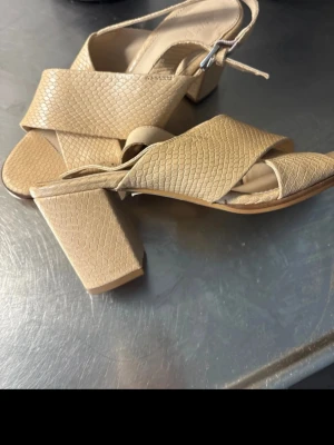 Beige sandaler med blockklack - Säljer ett par beige sandaler med breda korslagda remmar och struktur i ormskinnsmönster. Skorna har öppen tå, justerbar rem med spänne och stabil blockklack. Perfekta för dig som vill ha en trendig och bekväm look.