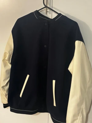 Svart bomberjacka med vita ärmar - Snygg svart bomberjacka i klassisk varsity-stil med kontrasterande vita ärmar och detaljer. Jackan har tryckknappar framtill och två vita fickor. Materialet ser ut att vara en mix av tyg och konstläder. Perfekt för en trendig streetwear-look.