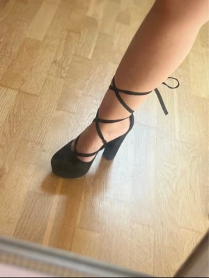 Svarta platåpumps med snörning - Säljer ett par svarta pumps med hög platå och chunky klack. Skorna har rund tå och tunna snören som lindas runt ankeln för en trendig look. Materialet ser ut att vara mockaimitation. Perfekta för dig som vill sticka ut!