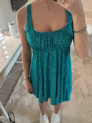 Turkos blommig kortklänning - Söt turkos kortklänning med små vita blommor och breda axelband. Klänningen har en luftig passform och lätt rynk vid bysten för en avslappnad look. Perfekt för varma dagar och enkel att styla med smycken eller sneakers.