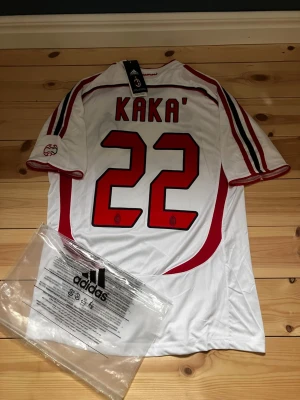 AC Milan Kaka' 22 Adidas matchtröja - Vit AC Milan matchtröja från Adidas med Kaka' och nummer 22 på ryggen. Tröjan har röda och svarta detaljer, Champions League-märke, Bwin-logga på bröstet och klubbmärke. Korta ärmar och sportig passform, perfekt för fotbollsfans.