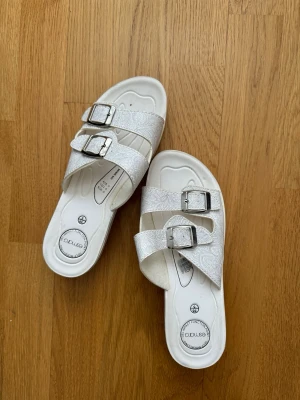 Vita sandaler med dubbla spännen - Snygga vita sandaler med två breda remmar och silverfärgade spännen. Remmarna har ett diskret mönster som ger extra stil. Fotbädden är formad för komfort och sulan är i slitstarkt material. Perfekta för sommaren och lätta att matcha.