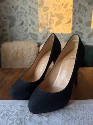 Svarta pumps från Christian Louboutin - Säljer ett par klassiska svarta pumps från Christian Louboutin med ikonisk röd undersida. Skorna har rundad tå och höga stilettklackar. Ytmaterialet är mörk grå textil och insidan är ljus läder. Har endast testats men passade mig tyvärr inte. Skorna passar till alla outfits, från kostymbyxor till klänning! Kontakta gärna kring funderingar💕