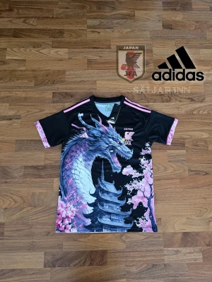 Japan Fotbollströja - Japan Dragon Fotbollströja – Limited Edition från Adidas. Storlek S (Slim Fit). Svart med rosa detaljer, tryck av japansk drake, pagoda och körsbärsblommor. Officiellt licensierad av JFA. 100 % polyester (AEROREADY) – lätt och snabbtorkande. Tagg avklippt. Perfekt för fans, cosplay eller streetwear.  📦 Snabb leverans – skickas samma dag! Kontakta mig vid intresse! 📌