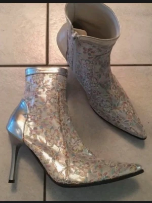 Silvriga stövletter med blommönster - Unika stövletter med spetsig tå och hög klack i metallic silver. Skorna har ett vackert blommönster i pastellfärger och dragkedja på sidan. Materialet är glansigt och ger en lyxig känsla. Perfekt för dig som vill sticka ut med din stil.