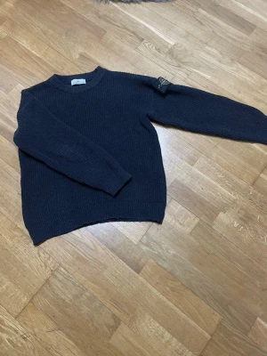 Mörkblå stickad tröja Stone Island - Säljer en mörkblå stickad tröja från Stone Island med klassisk rund hals och ribbstickade muddar. Tröjan har en diskret patch på ärmen och är gjord i ett tjockare material som passar perfekt för kyligare dagar.