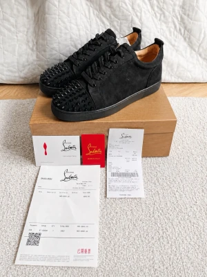 Svarta Louboutin sneakers med nitar - Svarta sneakers från Christian Louboutin i mocka med svarta nitar på tån. Skorna har lågt skaft, svarta skosnören och en diskret svart sula. Insidan är ljusbeige och materialet känns lyxigt och stilrent. Perfekta för dig som vill sticka ut med en edgy vibe.