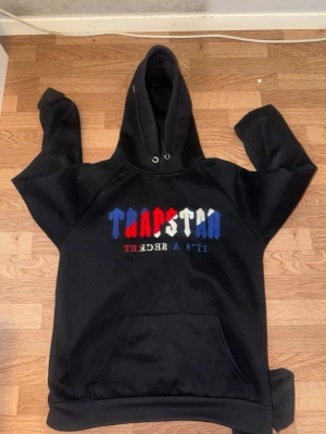 Svart Trapstar hoodie med tryck - Säljer en svart hoodie från Trapstar med färgglatt tryck på bröstet i rött, vitt och blått där det står 'TRAPSTAR' och 'IT'S A SECRET'. Hoodien har stor huva, känguruficka och ribbade muddar. Perfekt för dig som gillar streetwear och vill sticka ut.