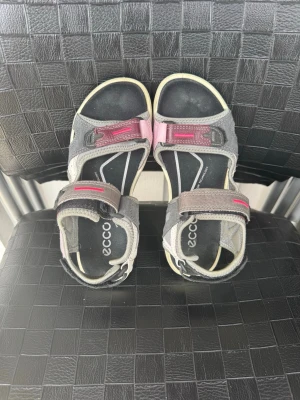 Grå och rosa sandaler från ECCO - Snygga och sportiga sandaler från ECCO i grått och rosa med svarta detaljer. De har justerbara kardborreband och en robust sula som passar perfekt för äventyr. Tillverkade i slitstarkt syntetmaterial och textil för maximal komfort.