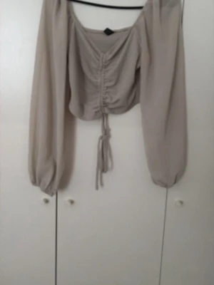 Beige omlottopp med snörning - Trendig beige topp med långa, puffiga ärmar och snygg snörning framtill. Toppen har omlottdesign och croppad passform som ger en modern look. Perfekt att styla med höga jeans eller kjol.