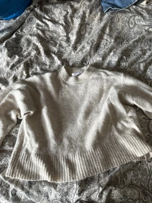 Beige stickad tröja med ribbad kant - Mysig beige stickad tröja med rund hals och bred ribbad kant nedtill. Tröjan har en avslappnad passform och långa ärmar, perfekt för lager på lager. Materialet ser ut att vara mjukt och skönt, troligen en ull- eller bomullsblandning.