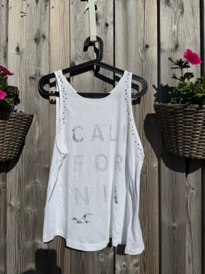 Vit linnetopp från Hollister - Säljer en vit linnetopp från Hollister med texten 'California' framtill och små silverfärgade nitar längs axlarna. Toppen är ärmlös och har en avslappnad passform, perfekt för varma dagar.