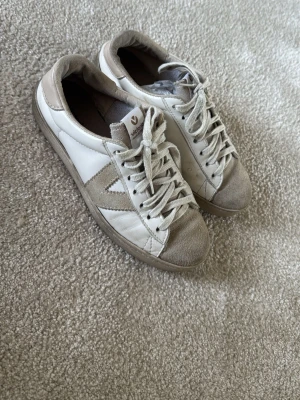 Vita och beige sneakers från Victoria - Snygga sneakers från Victoria i vitt och beige med mockadetaljer på tå och häl. Klassisk låg modell med snörning och sula i gummi. Perfekta för en avslappnad och trendig stil.