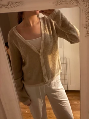 Beige kofta med knappar - Mysig beige kofta med v-ringning och långa ärmar. Koftan har ribbade muddar och stängs med fyra vita knappar framtill. Perfekt att slänga över ett linne eller t-shirt för en avslappnad look.