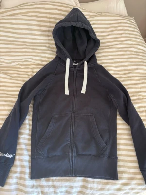 Mörkblå hoodie från Russemerch - Mörkblå hoodie med dragkedja från Russemerch. Har vit dragsko i huvan, två stora fickor framtill och ribbade muddar vid ärmslut och nederkant. Broderad Russedress-logga på ena ärmen. Perfekt för chill dagar och streetwear-stil.