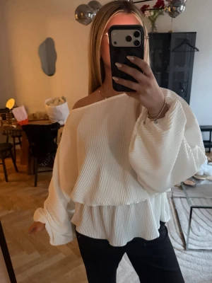 Vit plisserad offshoulder-blus - Trendig vit blus med plisserat tyg och offshoulder-design. Blusen har vida, långa ärmar och en lös, flowy passform med volang nedtill. Perfekt för dig som gillar en avslappnad men ändå chic stil. Materialet känns lätt och luftigt.