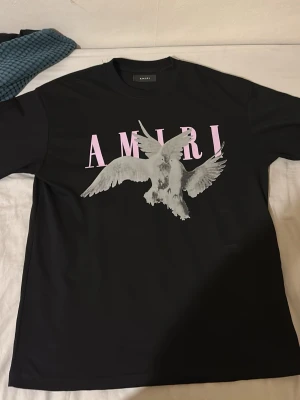 Svart AMIRI t-shirt med fågeltryck - Snygg svart t-shirt från AMIRI med stort grafiskt tryck av en fågel och rosa AMIRI-text på bröstet. Klassisk rund hals och korta ärmar. Perfekt för dig som vill ha en statement tee med exklusiv känsla.